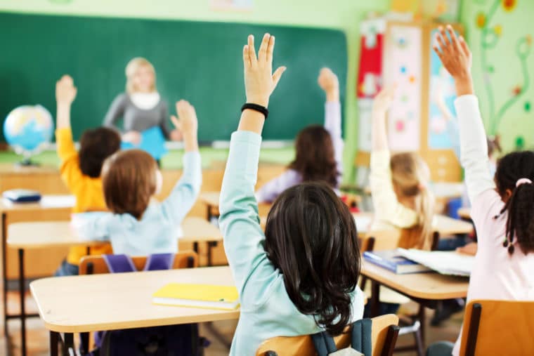 kids raising hands in a classroom point of view is from behind