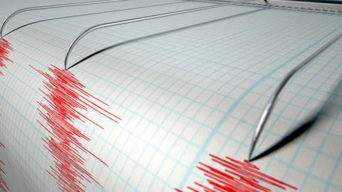 A closeup of a seismograph machine needle drawing a red line on graph paper depicting seismic and earthquake activity on an isolated white background
