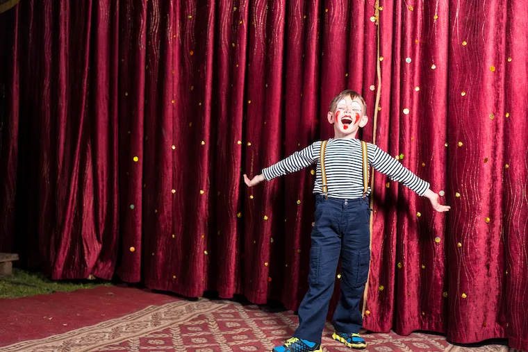 Adorable little boy singing on stage during a play standing with outstretched arms in his costume and makeup in front of the burgundy colored curtain