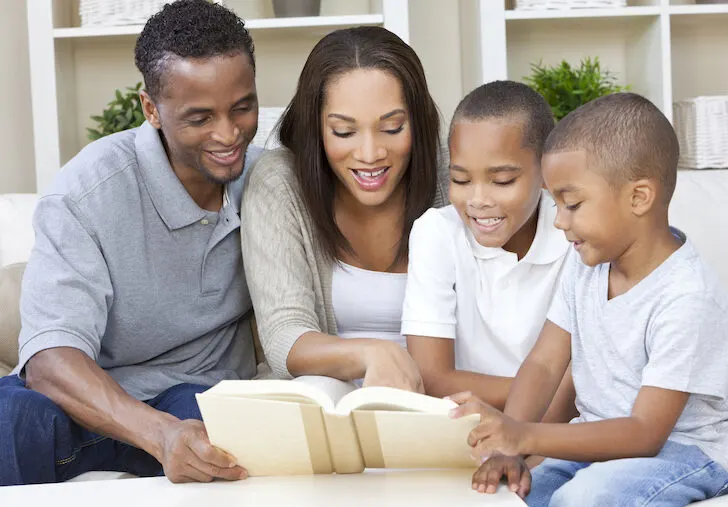 A happy African American man, woman and boys, father, mother and two sons, family sitting together at home reading a book